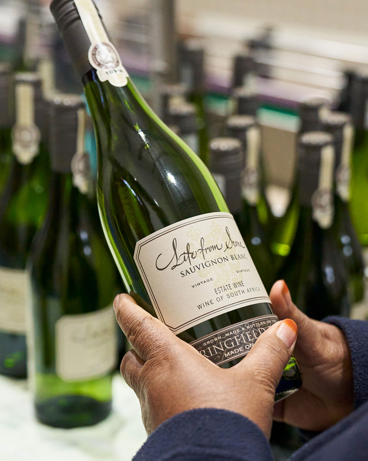 Eine Person hält eine Flasche Sauvignon Blanc mit der Aufschrift Solefrom Vine vor mehreren ähnlichen grünen Weinflaschen auf einem Tisch.