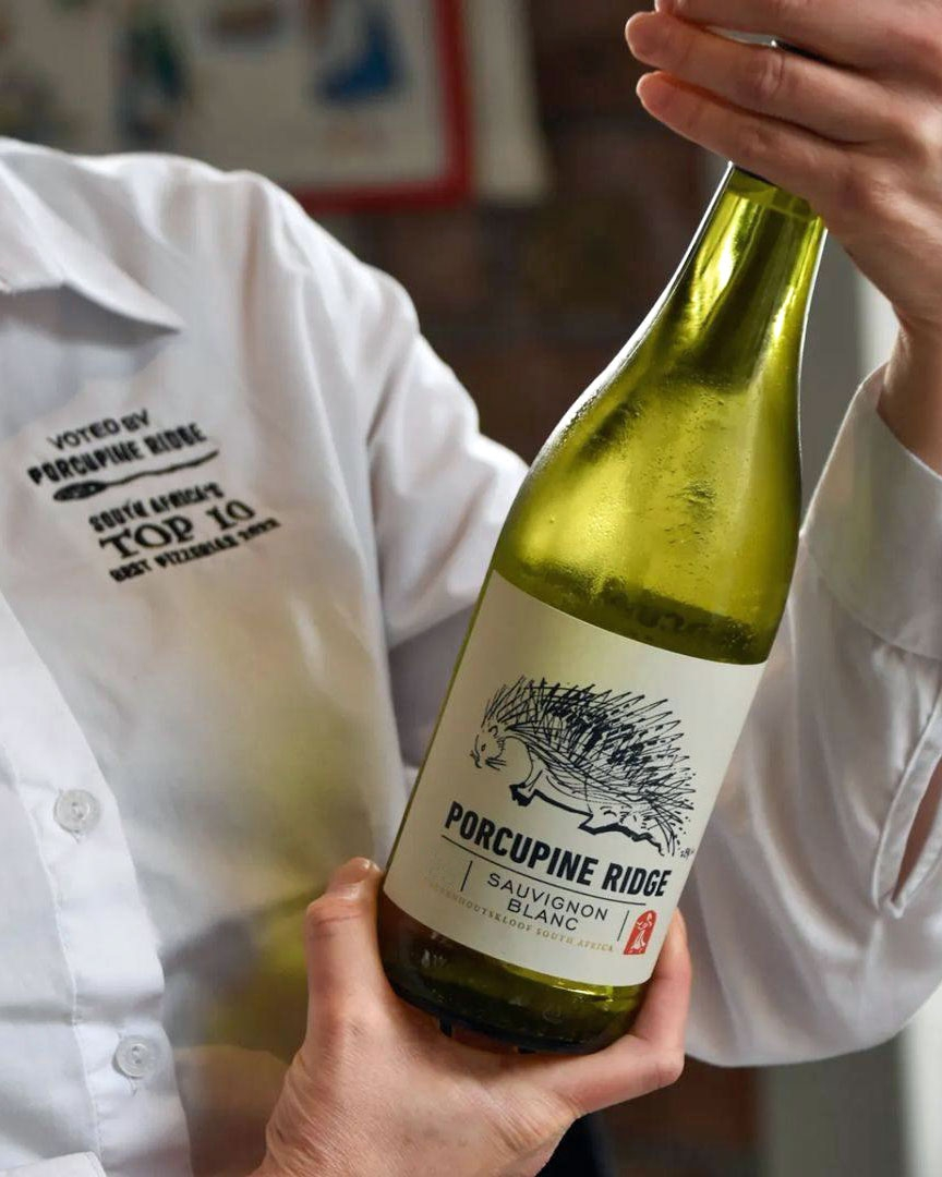 Eine Person, die ein weißes Hemd trägt, hält eine Flasche Porcupine Ridge Sauvignon Blanc Wein, auf dessen Etikett ein Stachelschwein abgebildet ist. Auf dem Hemd ist ein Text auf der Brust aufgedruckt.