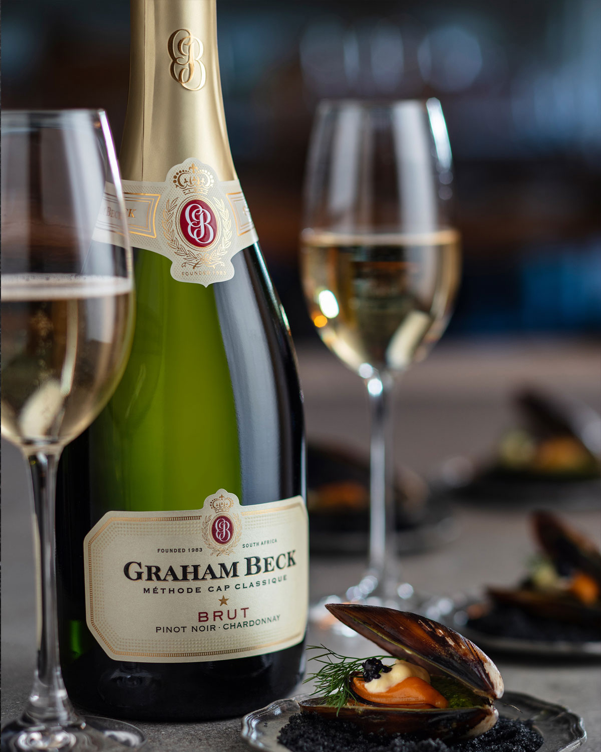 Eine Flasche Graham Beck Brut Sekt steht neben zwei gefüllten Weingläsern. Im Vordergrund wird auf einem schwarzen Teller eine Gourmet-Vorspeise mit Muschel und Beilage serviert. Das Ambiente wirkt elegant und raffiniert.