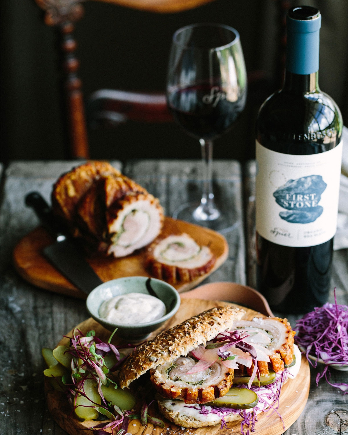 Ein rustikaler Tisch mit einem Sandwich, gefüllt mit Aufschnitt, Essiggurken und Rotkohl, daneben eine Schüssel mit Soße. Im Hintergrund sind ein Glas und eine Flasche Rotwein sowie ein Brotlaib zu sehen.