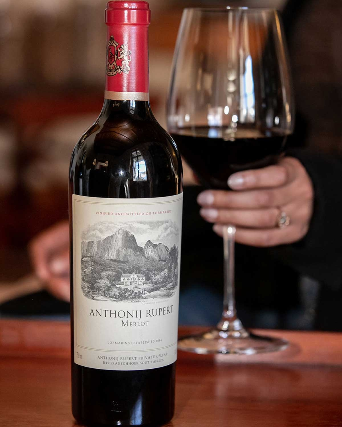 Eine Flasche Anthonij Rupert Merlot mit beigefarbenem Etikett, auf dem Berge und ein Weinberg abgebildet sind, und rotem Verschluss mit goldenem Wappen.