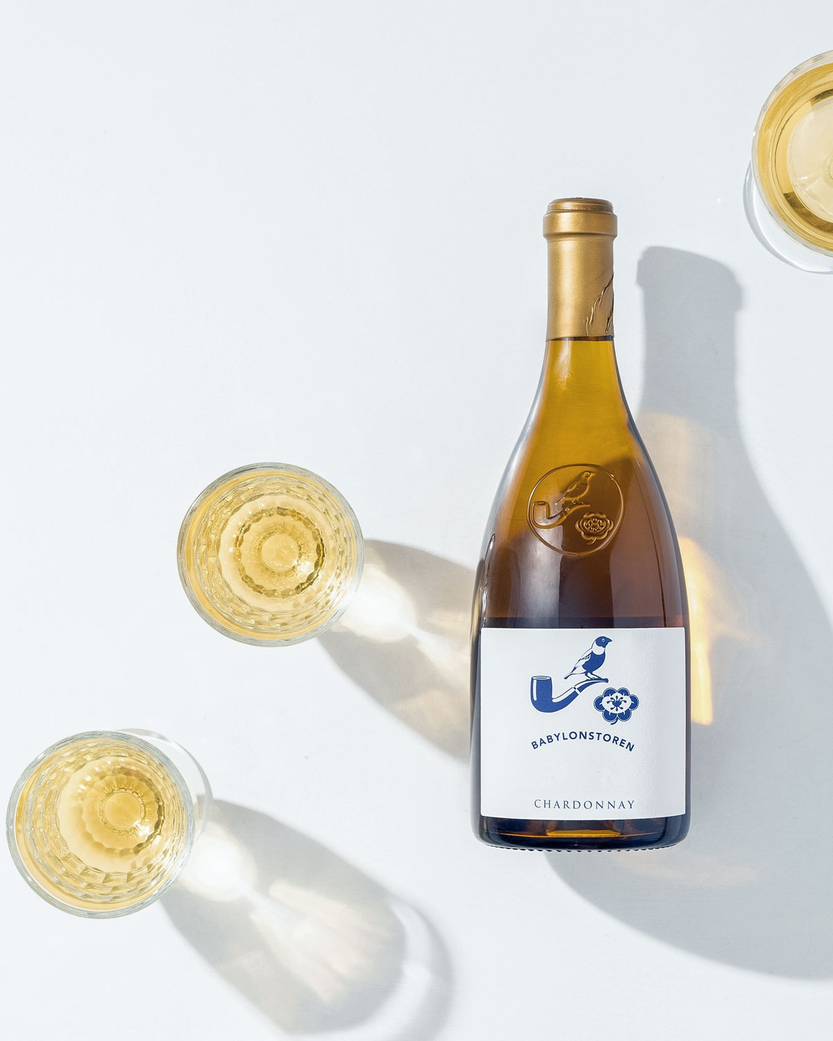 Eine Flasche Babylonstoren Chardonnay steht auf einer weißen Fläche neben drei gefüllten Weingläsern und wirft weiche Schatten in das Licht.