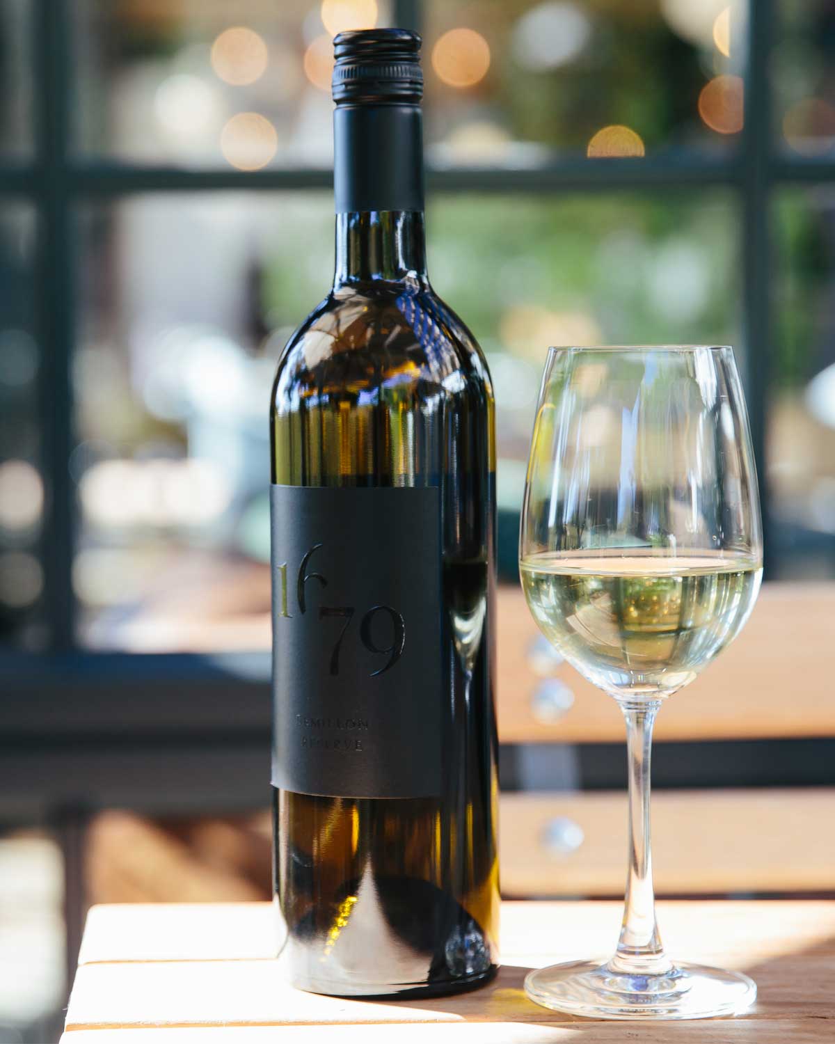 Eine Weißweinflasche mit der Aufschrift 1679 steht neben einem gefüllten Weinglas auf einem Holztisch, mit einem unscharfen Fenster und einer Szene im Freien im Hintergrund.