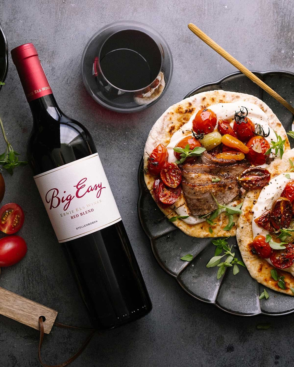 Eine Flasche Big Easy Rotwein liegt neben einem Glas Wein und zwei Fladenbroten mit gegrilltem Fleisch, Tomaten und Kräutern auf einem dunklen Teller, umgeben von frischen Tomaten auf einer grauen Fläche.