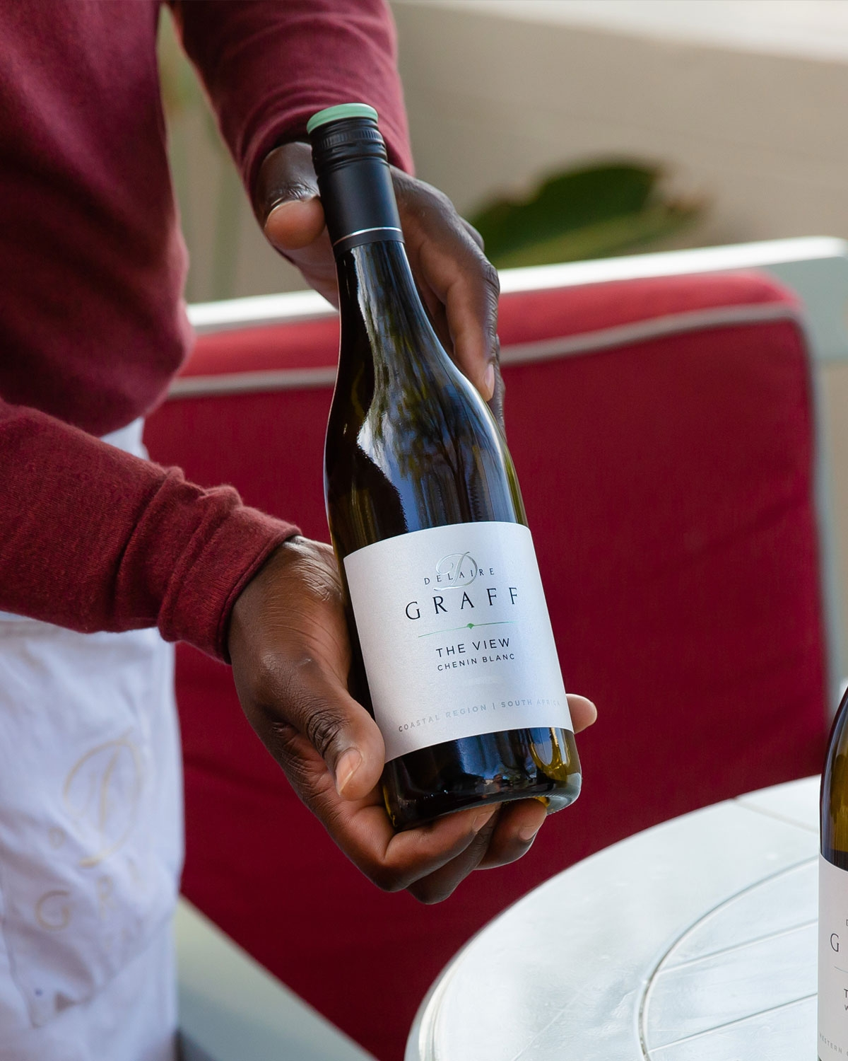 Eine Person in einem roten, langärmeligen Hemd hält eine Flasche Delaire Graff The View Chenin Blanc an einem Tisch mit rot gepolsterten Sitzgelegenheiten im Hintergrund.