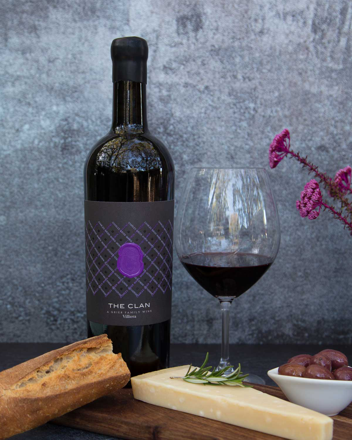 Eine Flasche Rotwein mit der Aufschrift "The Clan" steht neben einem Glas Rotwein, einem Laib Brot, einem Stück Käse mit Rosmarin, einer kleinen Schale mit Oliven und violetten Blumen, alles auf einem strukturierten grauen Hintergrund.