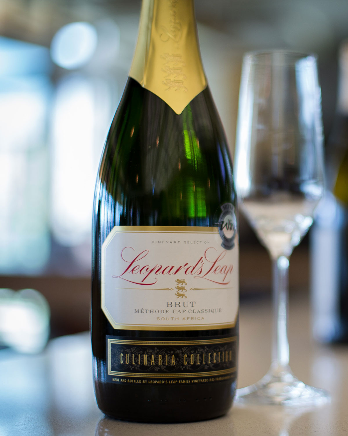 Eine Nahaufnahme einer Leopard's Leap Brut Schaumweinflasche aus Südafrika mit der Aufschrift Méthode Cap Classique steht neben einem leeren Weinglas auf einer spiegelnden Oberfläche.