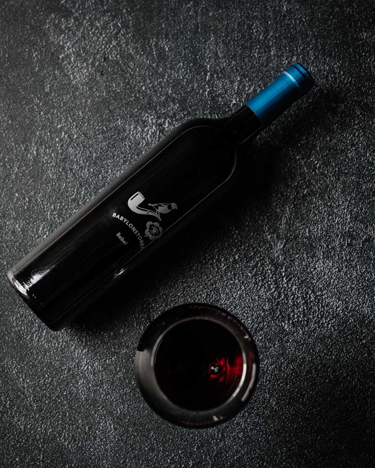 Eine große Weinflasche aus dunklem Glas mit blauem Foliendach und der Aufschrift BABYLONSTOREN Babel mit einem weißen Logo, das einen Vogel auf einer Pfeife und eine Blume zeigt.