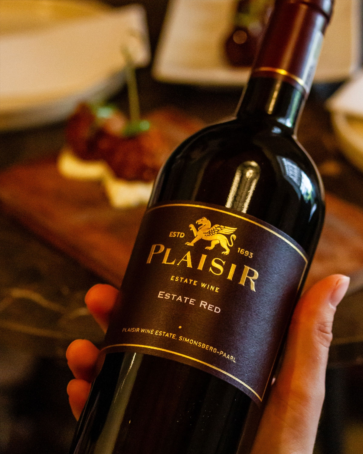 Eine dunkle Glasflasche des Plaisir Estate Rotweins mit einem braunen Etikett mit goldenen Akzenten, einem Löwenemblem und einem Text, der angibt, dass er vom 1693 gegründeten Plaisir Wine Estate in Simonsberg, Paarl, stammt.
