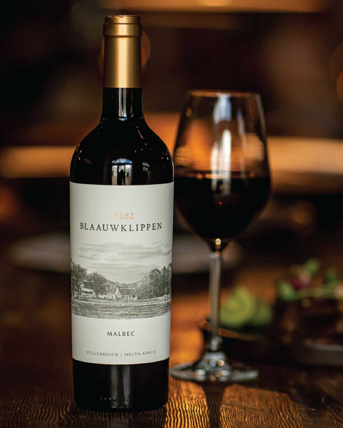 Eine Flasche Blaauwklippen Malbec-Wein und ein gefülltes Weinglas stehen auf einem Holztisch, mit verschwommenem Essen und warmer Beleuchtung im Hintergrund.