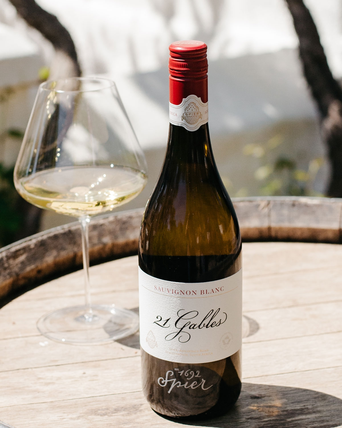 Eine Flasche des Weins Spier 21 Gables Sauvignon Blanc steht auf einem Holzfass neben einem Glas Weißwein, während das Sonnenlicht im Hintergrund Schatten wirft.