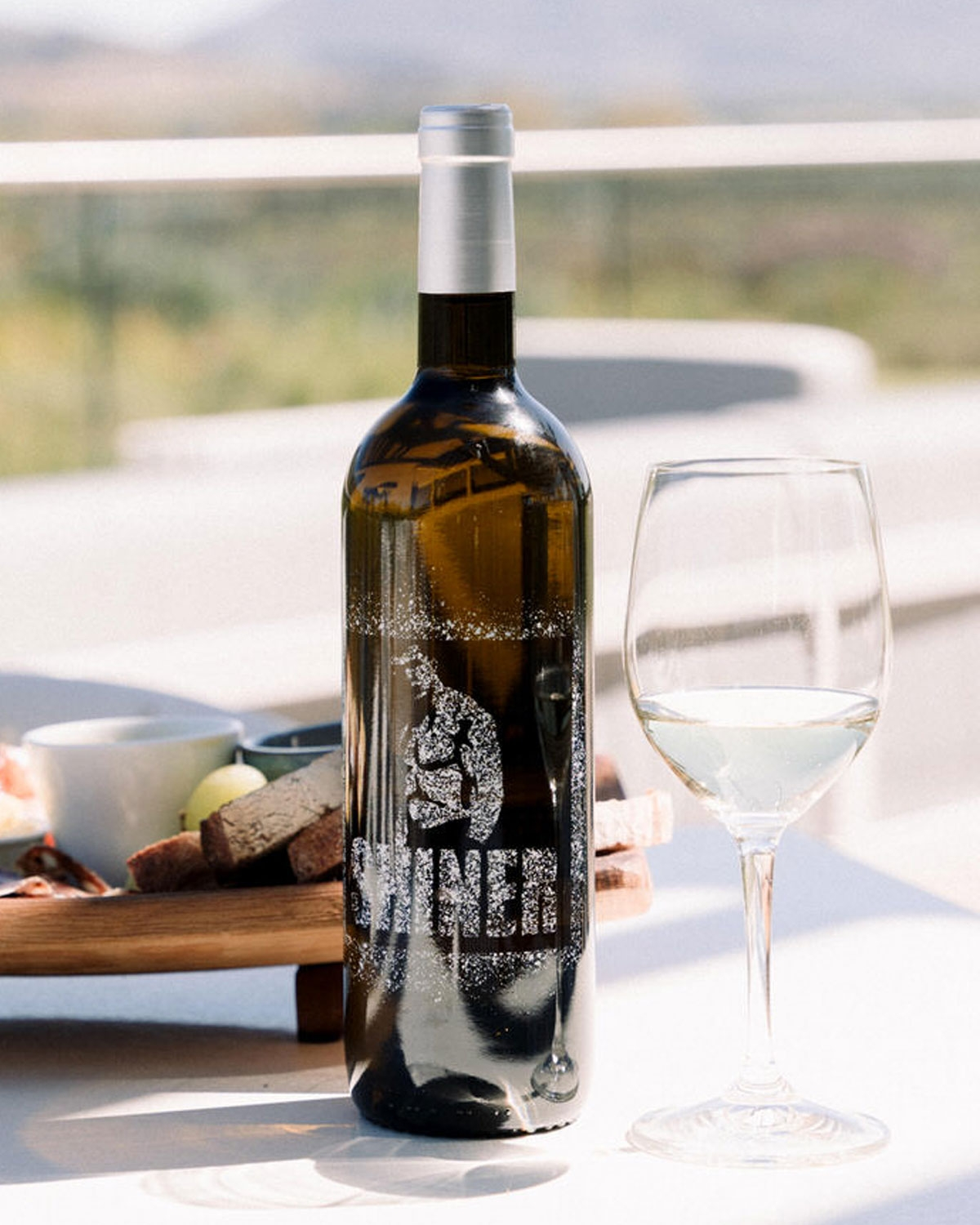 Eine Flasche Weißwein mit einem silbernen Verschluss steht neben einem gefüllten Weinglas auf einem sonnenbeschienenen Tisch. Im Hintergrund ist eine Holzplatte mit Brot, Käse und Obst zu sehen, dahinter ein Außenbereich.