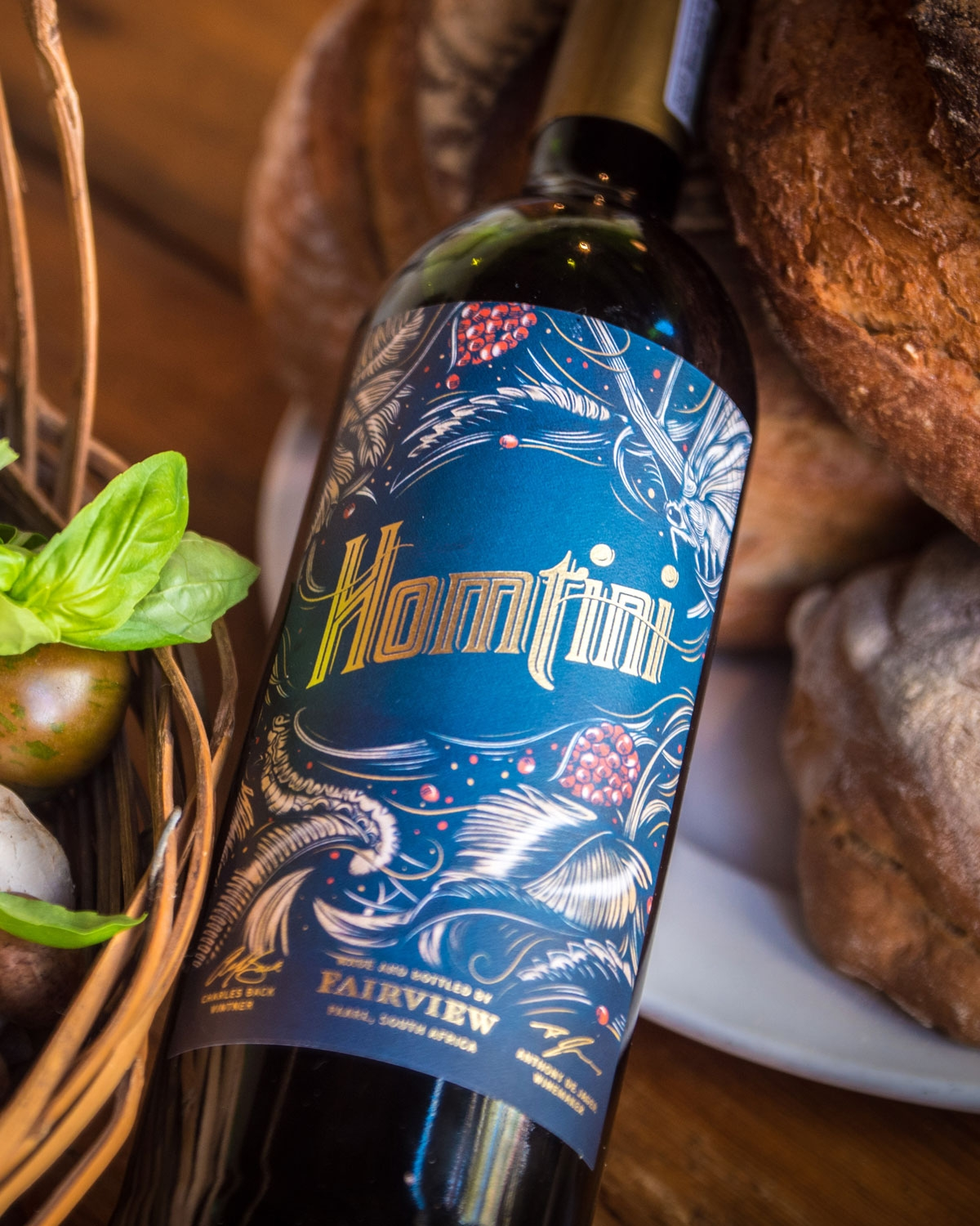 Eine Flasche Fairview Homtini-Wein mit verschnörkeltem Etikett liegt auf einem Tisch neben einem Korb mit Basilikum und Tomaten und einem Teller mit rustikalen Brotlaiben.