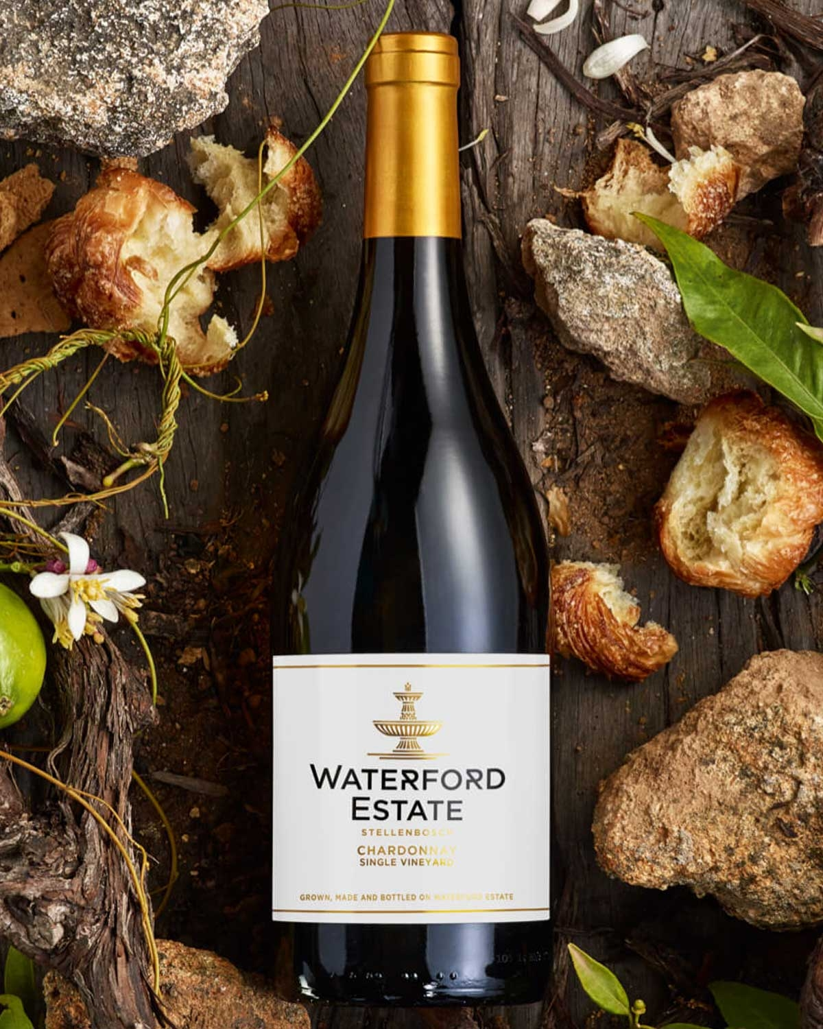 Eine Flasche Waterford Estate Chardonnay aus Stellenbosch mit einem Deckel aus Goldfolie und einem weißen Etikett mit goldenem und schwarzem Text, das aufrecht vor einem schlichten Hintergrund steht.