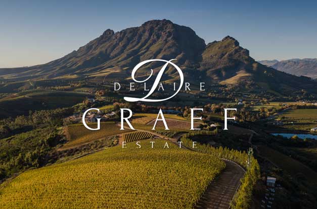 Luftaufnahme der Weinberge von Delaire Graff Estate mit einem gewundenen Pfad, üppigem Grün und einem großen Berg im Hintergrund unter einem klaren blauen Himmel. Der Text von Delaire Graff Estate ist in der Mitte überlagert.
