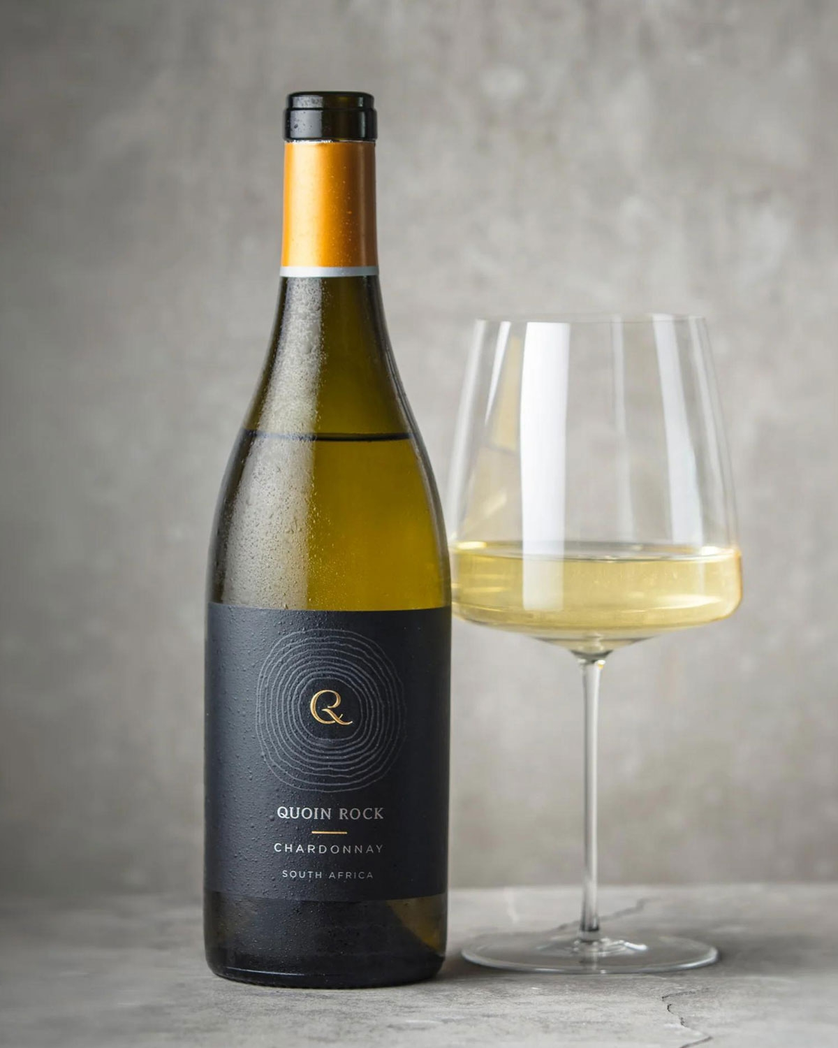 Eine Flasche Quoin Rock Chardonnay-Wein steht neben einem mit Weißwein gefüllten Glas, beide auf einer grauen Fläche mit neutralem grauem Hintergrund.