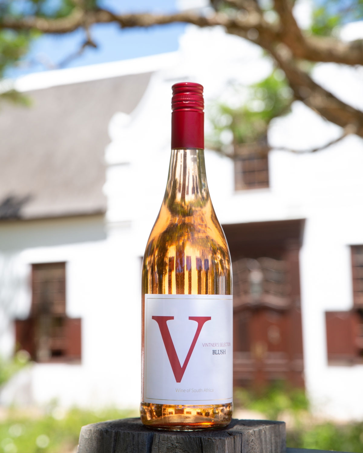 Eine Flasche Vintner's Selection Blush Wein steht auf einem Holzpfosten im Freien, mit einem weißen Gebäude und grünen Bäumen, die im Hintergrund verschwimmen.