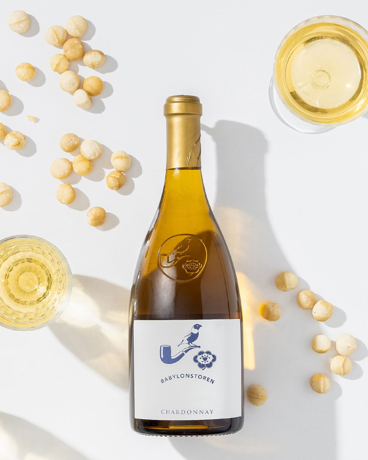 Eine Flasche Babylonstoren Chardonnay liegt auf einer weißen Fläche, umgeben von zwei Gläsern Weißwein und verstreuten Macadamianüssen. Die Flasche wirft einen deutlichen Schatten.