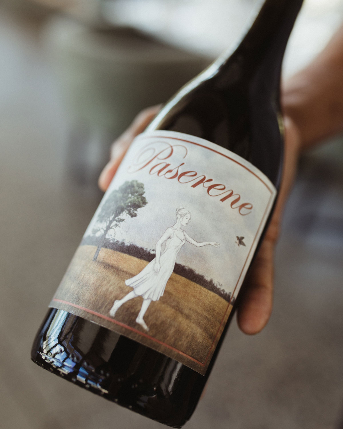 Eine Weinflasche mit einem blauen Wachssiegel und einem Etikett, das eine Frau in einem Kleid zeigt, die in einem Feld steht und auf einen Vogel zeigt, mit einem Baum im Hintergrund und der Aufschrift Pasarene.