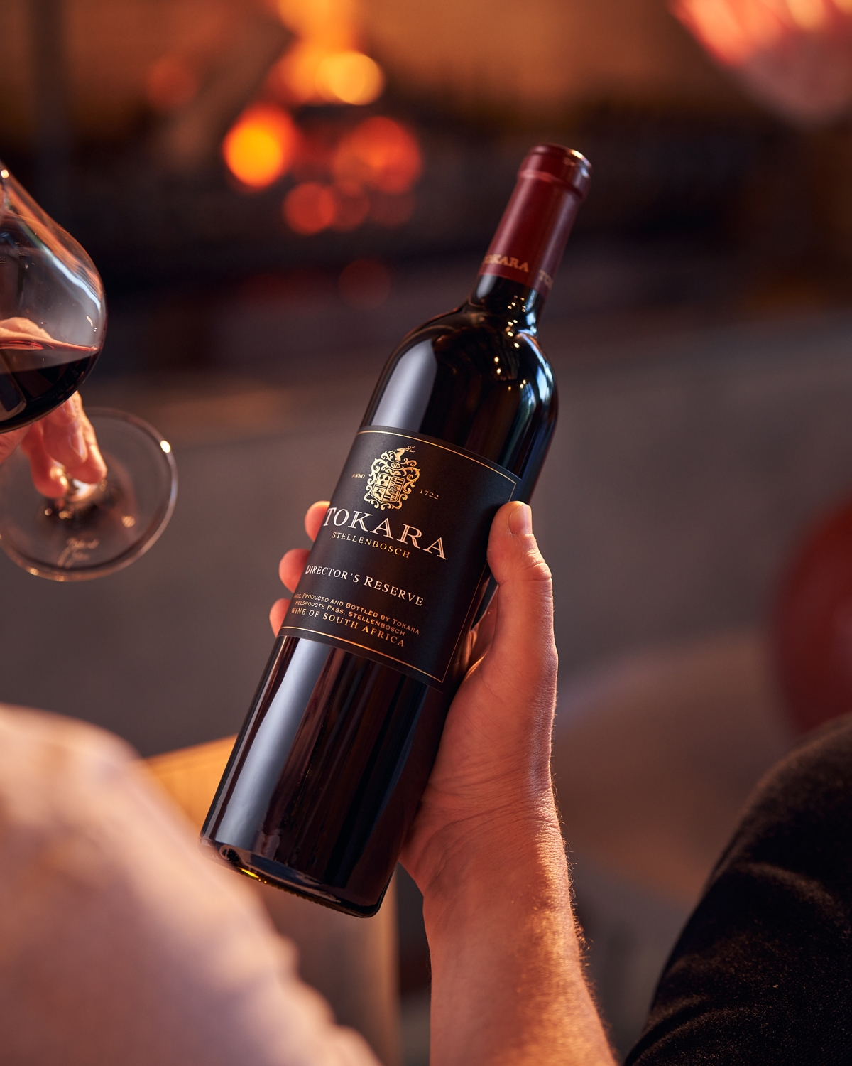 Eine Person hält eine Flasche Tokara Director's Reserve Rotwein in der Hand, während eine andere Person den Wein in einem Glas schwenkt; im Hintergrund leuchtet ein warmer Kamin und schafft eine gemütliche Atmosphäre.
