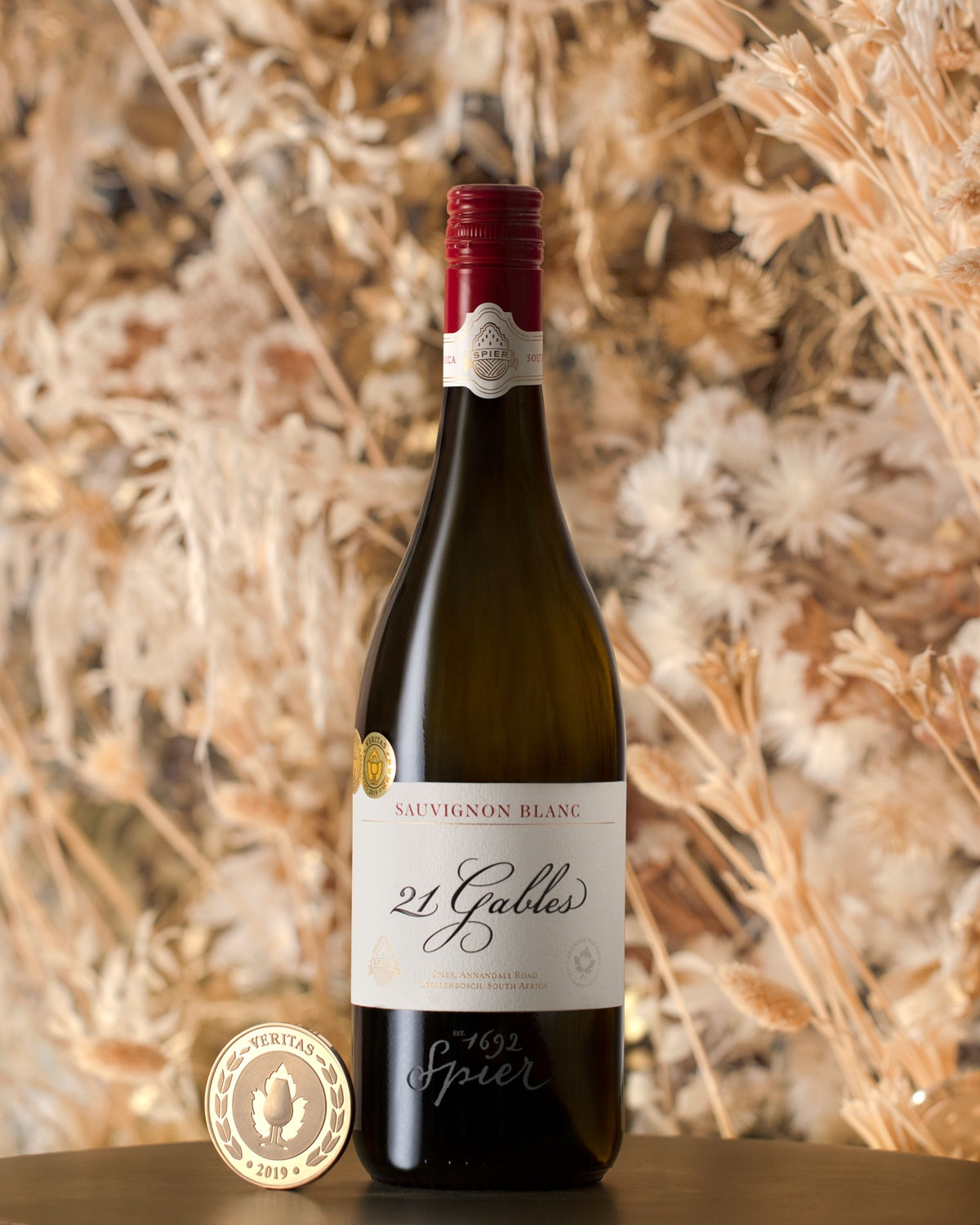 Eine Flasche Spier 21 Gables Sauvignon Blanc mit einem tiefroten Verschluss steht auf einem Tisch. Davor steht ein goldenes Medaillon der Veritas-Auszeichnung 2019, und den Hintergrund bilden getrocknete beigefarbene Blumendekorationen.