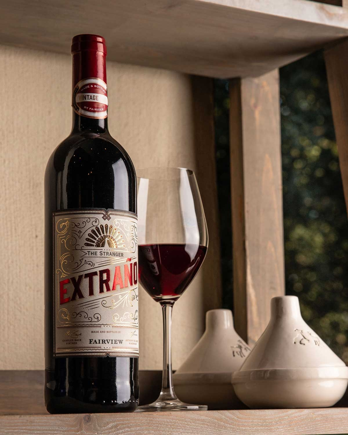 Eine Flasche Extraño-Rotwein steht neben einem gefüllten Weinglas auf einem Holzregal, mit zwei dekorativen Keramikbehältern im Hintergrund.