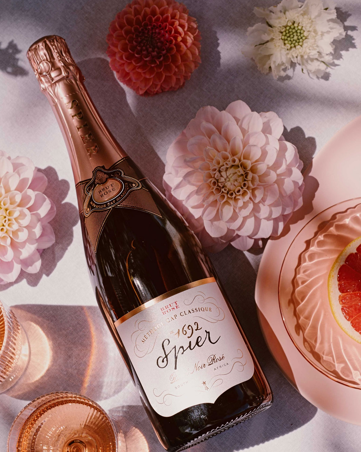 Eine Flasche Spier Méthode Cap Classique Brut Rosé steht auf einem Tisch mit rosafarbenen Blumen, rosafarbenen Gläsern und einer Schale mit einer Grapefruitscheibe, alles in einem sanften, natürlichen Licht.