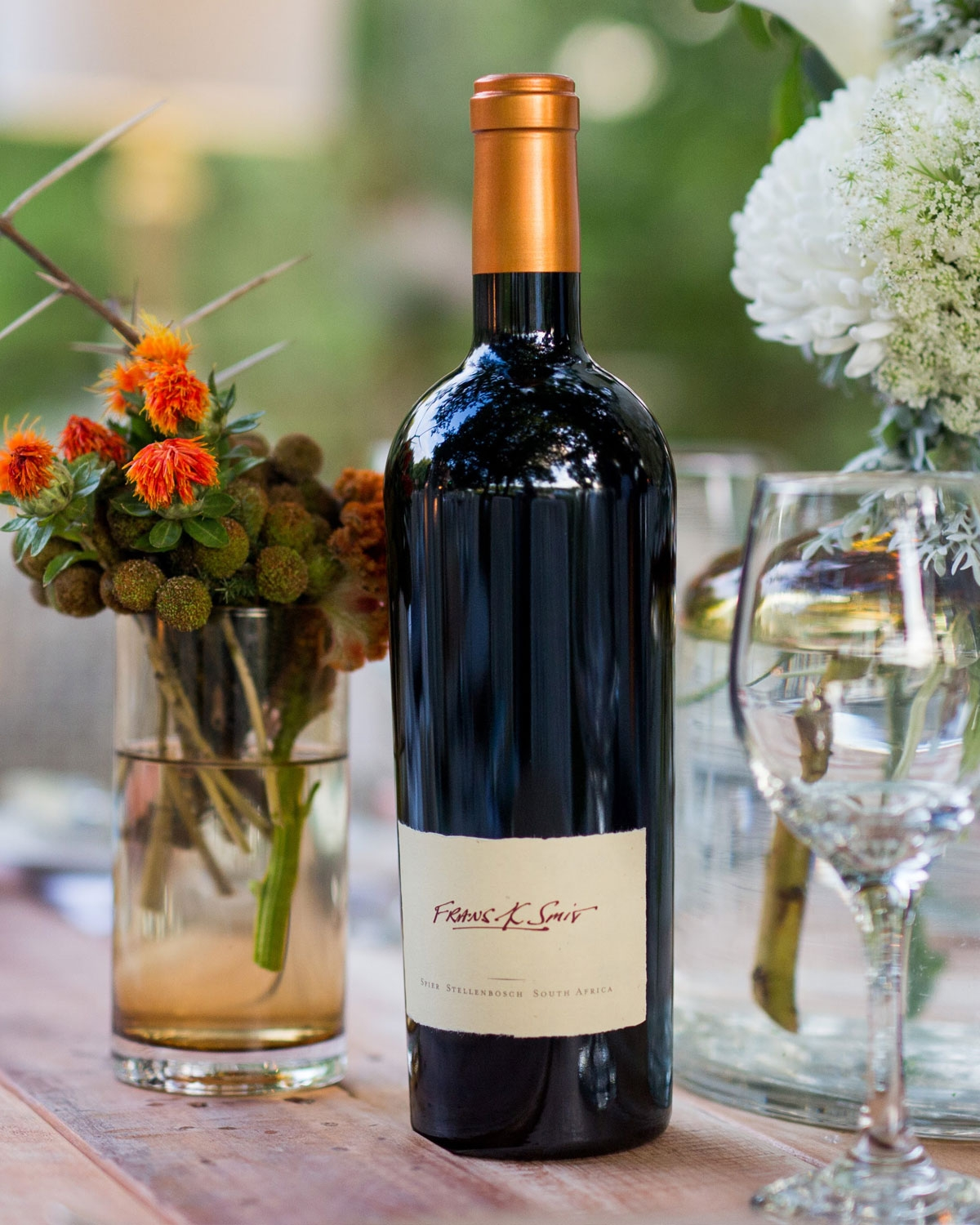 Eine Rotweinflasche mit kupferfarbenem Verschluss steht auf einem Holztisch, umgeben von Vasen mit orangefarbenen und weißen Blumen und leeren Weingläsern, mit einem unscharfen Hintergrund im Freien.