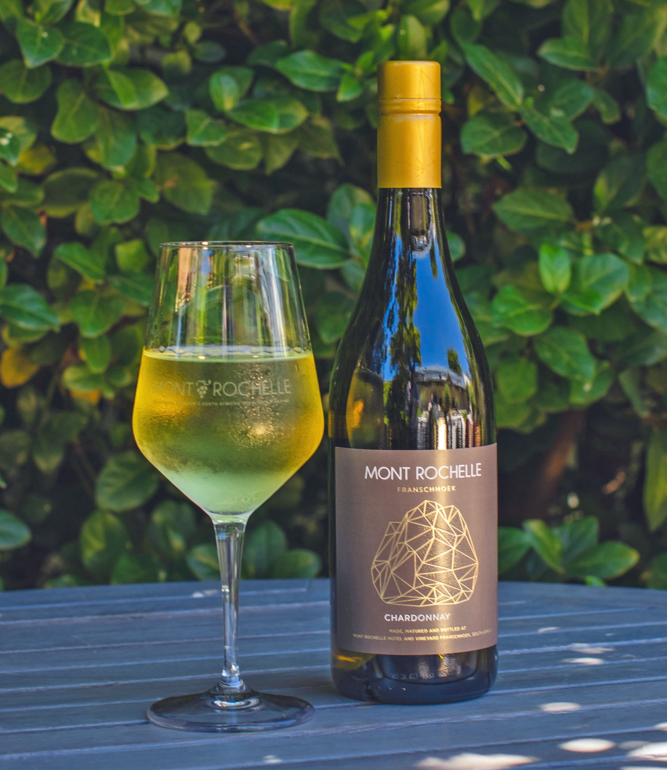Eine Flasche Mont Rochelle Franschhoek Chardonnay mit einem goldenen Schraubverschluss und einem dunkelbraunen Etikett mit einer geometrischen Illustration und goldener Schrift.