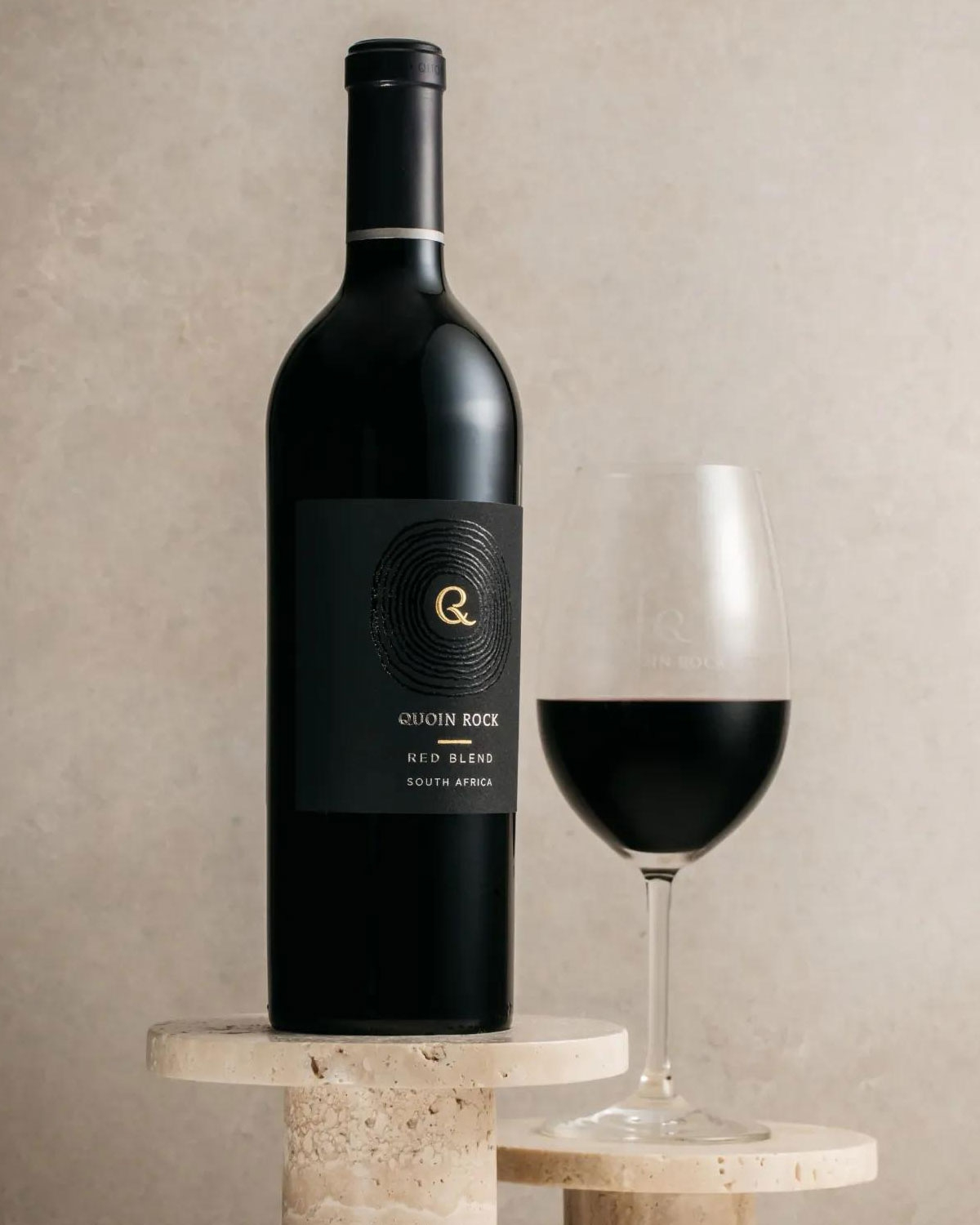 Eine Flasche Quoin Rock Red Blend Wein aus Südafrika steht neben einem Glas Rotwein auf einem cremefarbenen Steinpodest vor einem neutralen Hintergrund.