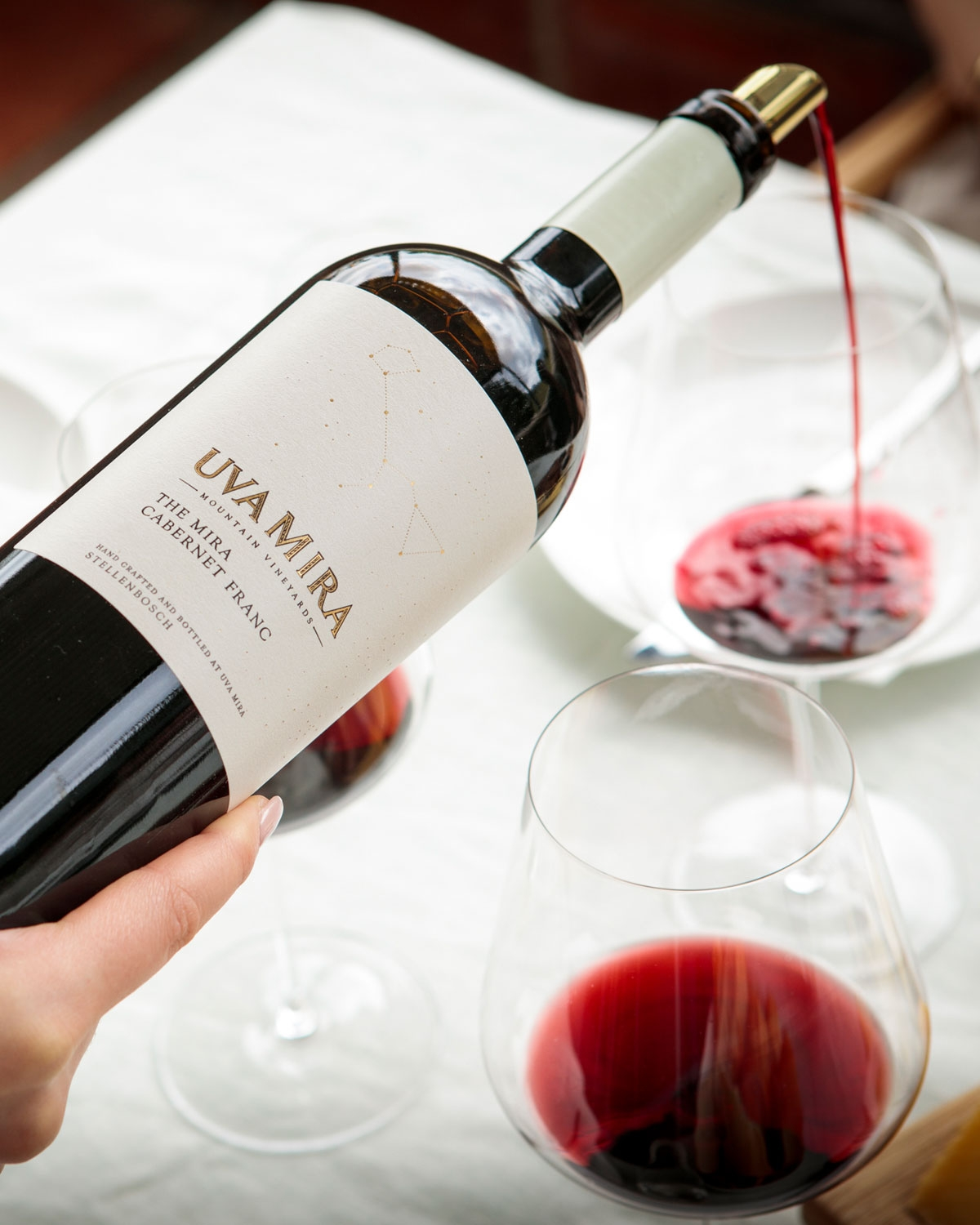 Eine Hand schenkt Rotwein aus einer Flasche mit der Aufschrift Uvamira The Mira Cabernet Franc in ein Weinglas auf einem hellen Tischtuch ein, daneben steht ein weiteres volles Glas Rotwein.