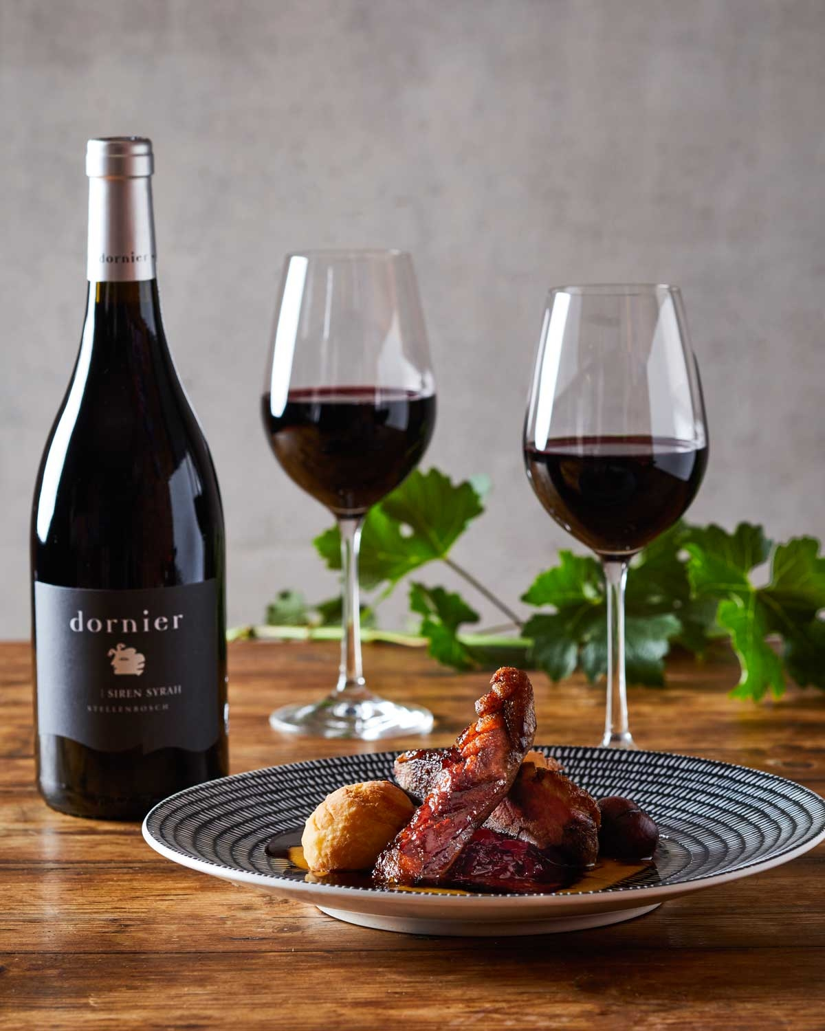 Eine dunkle Weinflasche mit silbernem Verschluss trägt ein schwarzes Etikett mit der Aufschrift "dornier Siren Syrah Stellenbosch" und ein kleines illustriertes Sirenenlogo auf der Vorderseite. Die Flasche steht aufrecht vor einem schlichten hellen Hinte