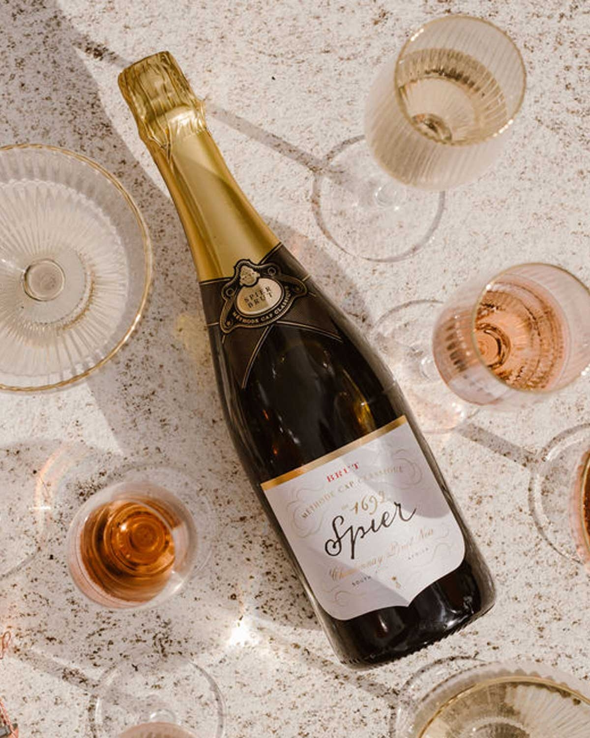 Eine Flasche Spier-Sekt liegt auf einer hellen Fläche, umgeben von verschiedenen Gläsern, die mit Rosé- und weißem Sekt gefüllt sind.
