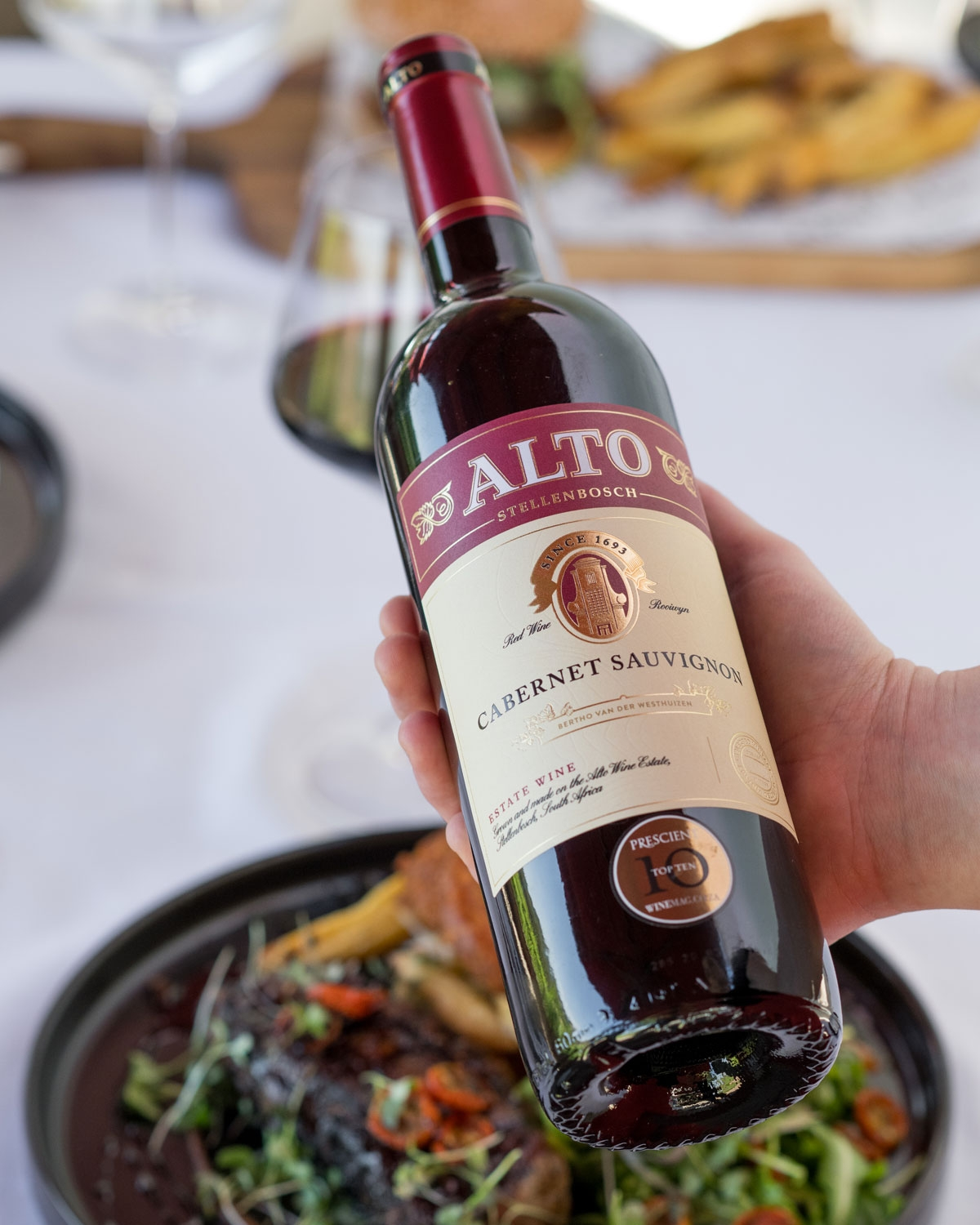 Eine Hand hält eine Flasche Alto Cabernet Sauvignon über einem gedeckten Tisch mit Speisen, darunter ein Fleischteller mit Grünzeug und ein Teller mit Pommes frites im Hintergrund.