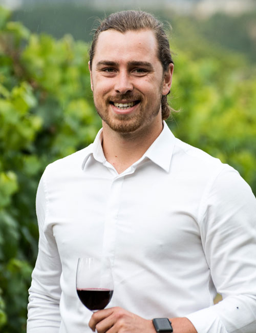 Ein Mann mit langem Haar und weißem Hemd steht im Freien und hält ein Glas Rotwein in der Hand. Grünes Laub füllt den unscharfen Hintergrund.