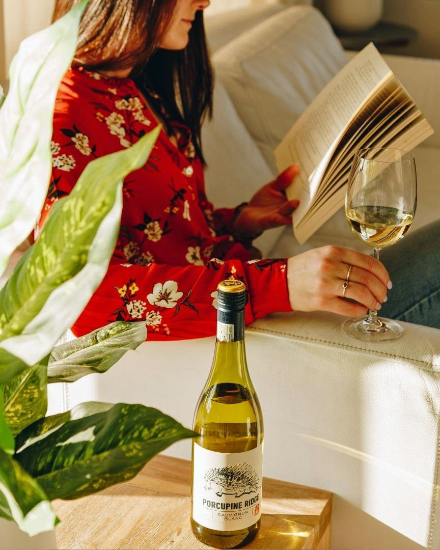Eine Frau in einer roten geblümten Bluse sitzt auf einer weißen Couch, liest ein Buch und hält ein Glas Weißwein in der Hand. Eine Flasche Porcupine Ridge-Wein steht auf einem Holztisch in der Nähe, während das Sonnenlicht einfällt und eine grüne 