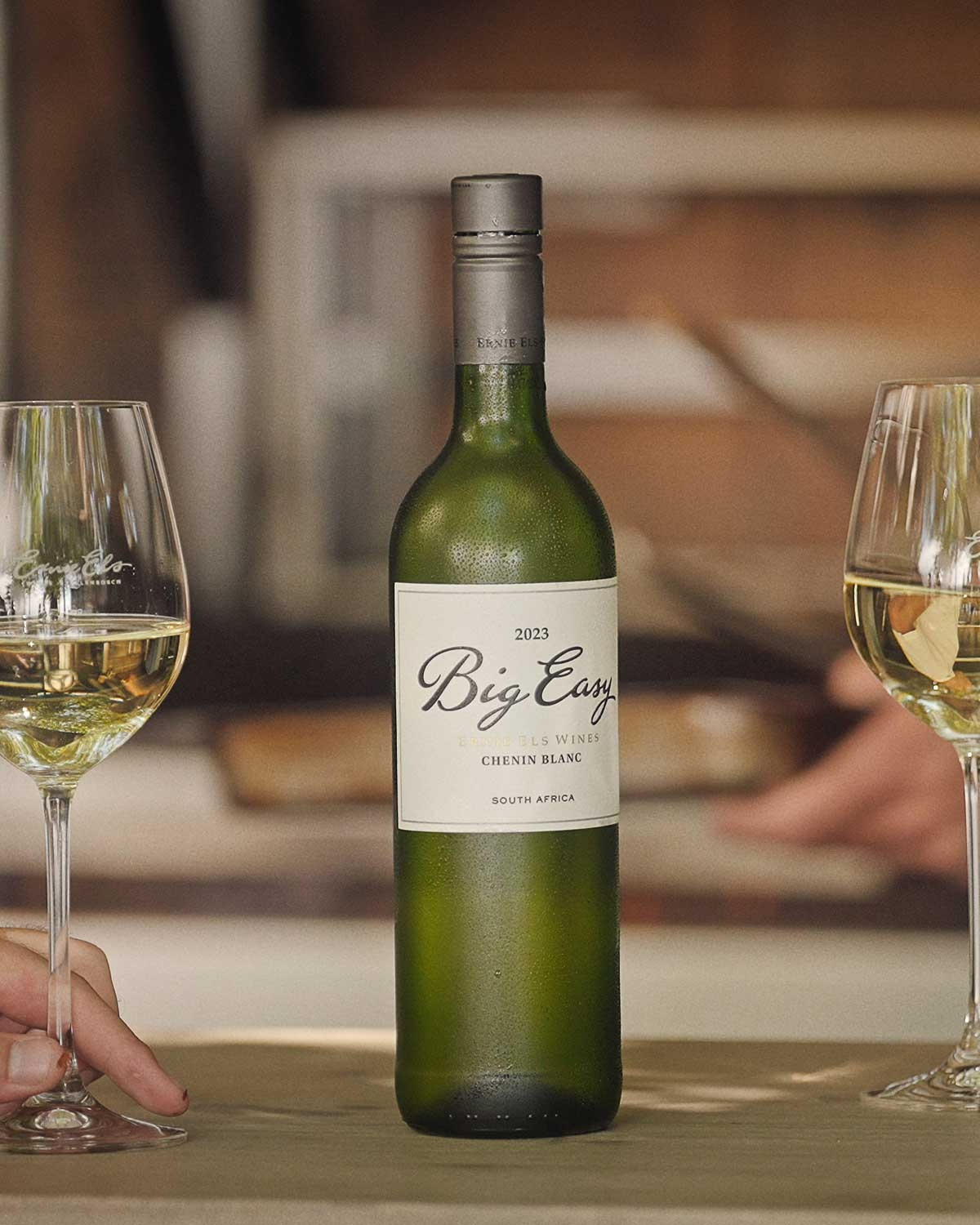 Eine gekühlte Flasche Big Easy 2023 Chenin Blanc aus Südafrika steht auf einem Tisch zwischen zwei gefüllten Weingläsern, und im Hintergrund sind zwei Hände zu sehen.