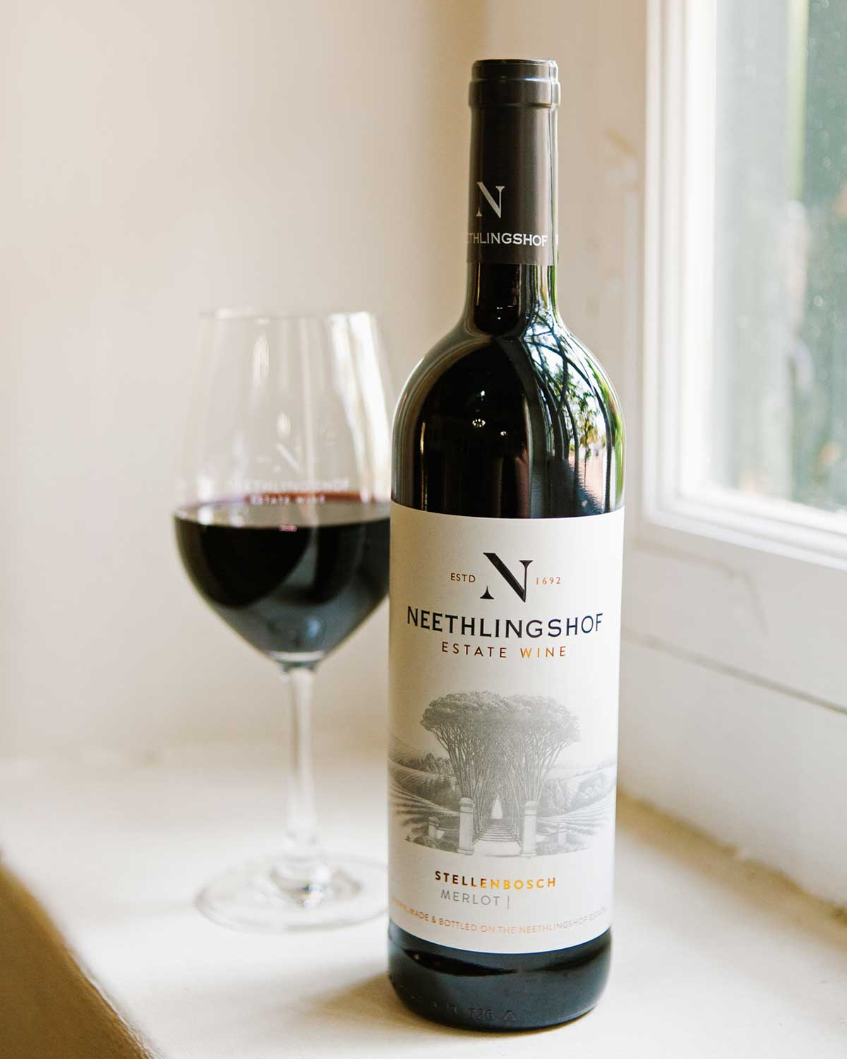 Eine Flasche des Neethlingshof Estate Wine Merlot aus Stellenbosch. Das Etikett zeigt eine Zeichnung eines von Bäumen gesäumten Weges und das Logo des Weinguts, mit elegantem, minimalistischem Design und goldenen Akzenten.