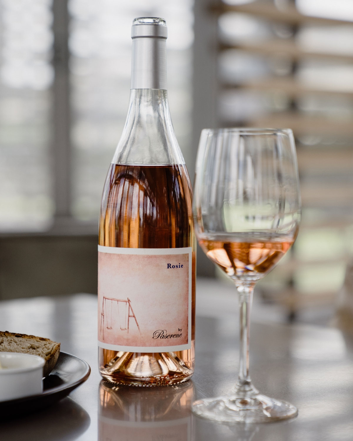 Eine Flasche Roséwein mit einem silbernen Verschluss. Das Etikett zeigt ein einfaches sepiafarbenes Bild einer leeren Schaukel und die Worte Rosie und by Pasarene. Der Wein hat eine leicht rosafarbene Tönung.